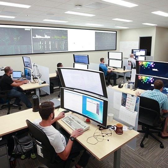 六名Viasat员工坐在2张大桌子前, 用于网络安全行动的堆叠监视器